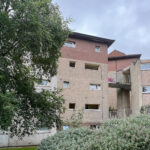 Mid-century apartment building with beige stucco and pink upper section, balconies and a sky-blue backdrop, partially obscured by green trees in the foreground