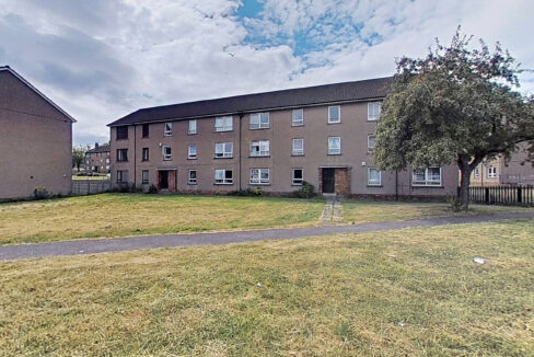 Three-story beige apartment building with many windows, a grassy foreground, and a large tree on the right under a cloudy blue sky.