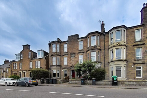 Row of brick townhouses along a street with parked cars and a row of trash bins in the foreground under a cloudy blue sky.