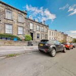 Row of stone-built terraced houses with white-framed windows along a sunny street; cars parked at the curb.