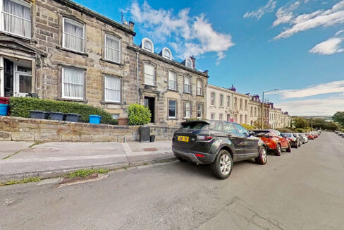 Row of stone-built terraced houses with white-framed windows along a sunny street; cars parked at the curb.