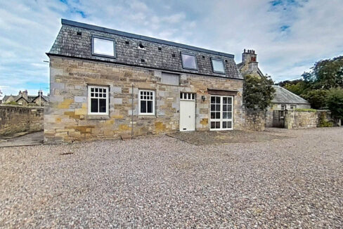 Historic stone house with a slate roof and white-framed windows, set in a gravel courtyard under a cloudy sky.