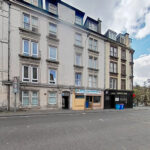 Row of multi-story beige buildings with ground-floor shops, including The Park Fish & Chicken Bar and Ross's Bar, under a blue sky with clouds