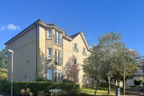 Three-story beige apartment building with stone accents, balconies, and a clear blue sky above.