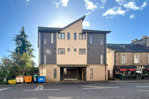 Modern four-story building with a ground-floor open garage, on a sunny street beside a bakery/c cafe and colorful waste bins on the left.