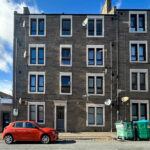 Three-story gray stone apartment building with white-trimmed windows, a red car parked on the street, and green dumpsters on the curb outside.
