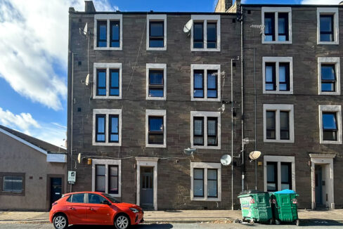Three-story gray stone apartment building with white-trimmed windows, a red car parked on the street, and green dumpsters on the curb outside.